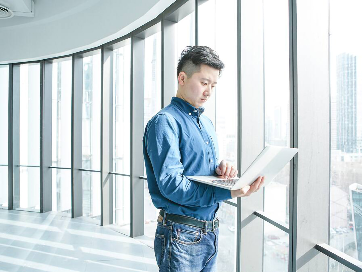 person looking at the laptop while standing near a window of a skyscraper